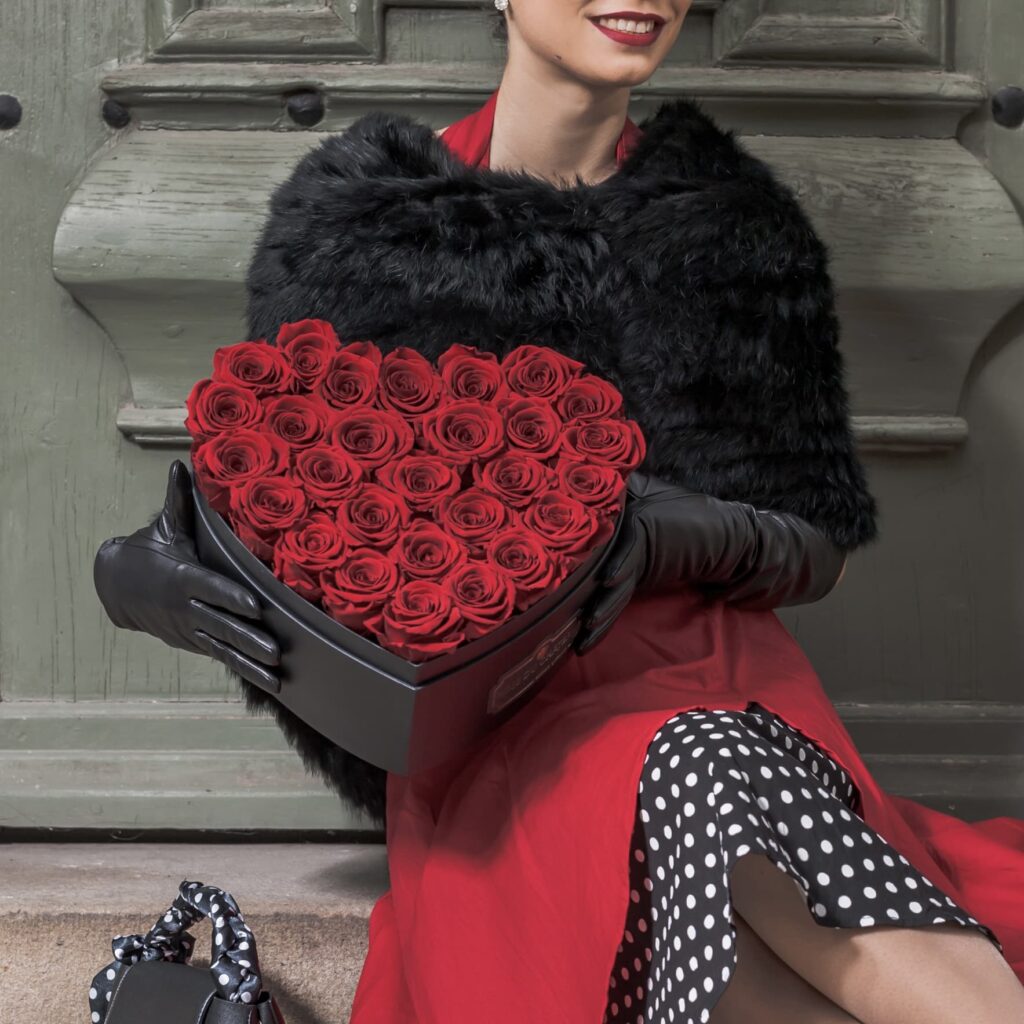 Jakie kwiaty na przeprosiny? Sprawdź wyjątkowy bukiet Flowerbox serce z rożami wiecznymi w kolorze czerwonym trzymany przez elegancką kobietę siedzącą na betonie